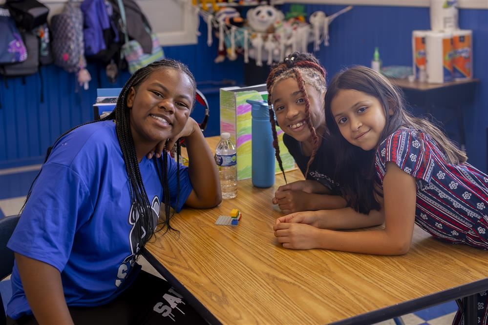 three girls smile at the camera while working on a group project. 